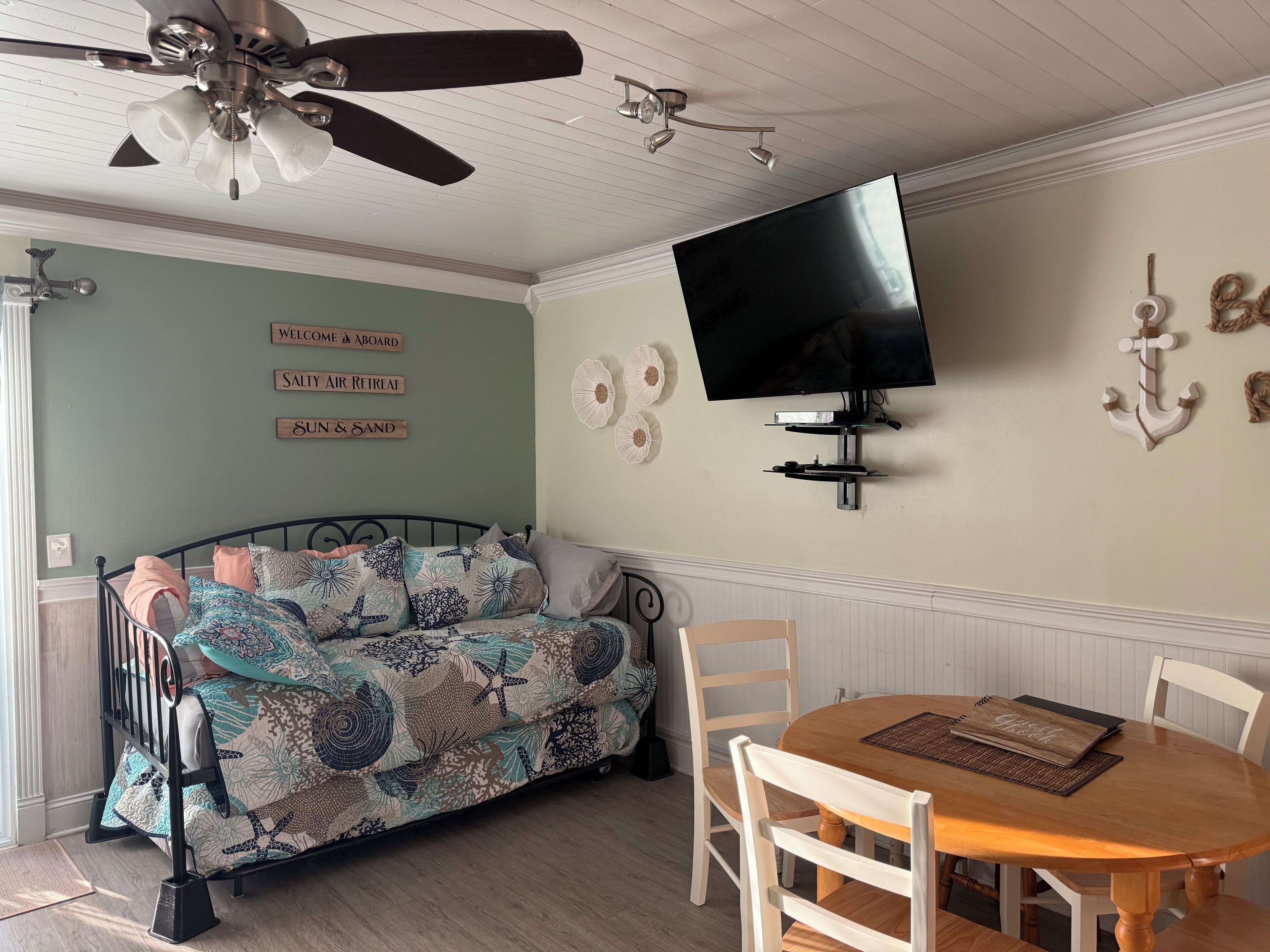 A room with a bed, a TV mounted on the wall, a ceiling fan, and a table with chairs.