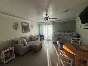 A living room with a sofa, ottoman, and a dining table with chairs. - Beautiful Condo In The Heart Of Carolina Beach (Carolina Beach)