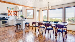 Une cuisine moderne avec un plancher en bois, des armoires blanches et un grand îlot central doté de tabourets. De grandes fenêtres offrent une vue sur la plage.