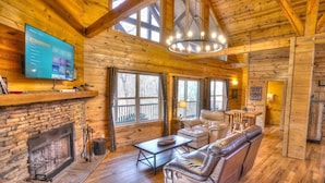 A cozy log cabin interior with a fireplace, leather seating, a dining area, and a large window overlooking a deck. - Allure Cabin – Family-Friendly Escape w/ Fire Pit, Creek & Near Helen! (Cleveland)