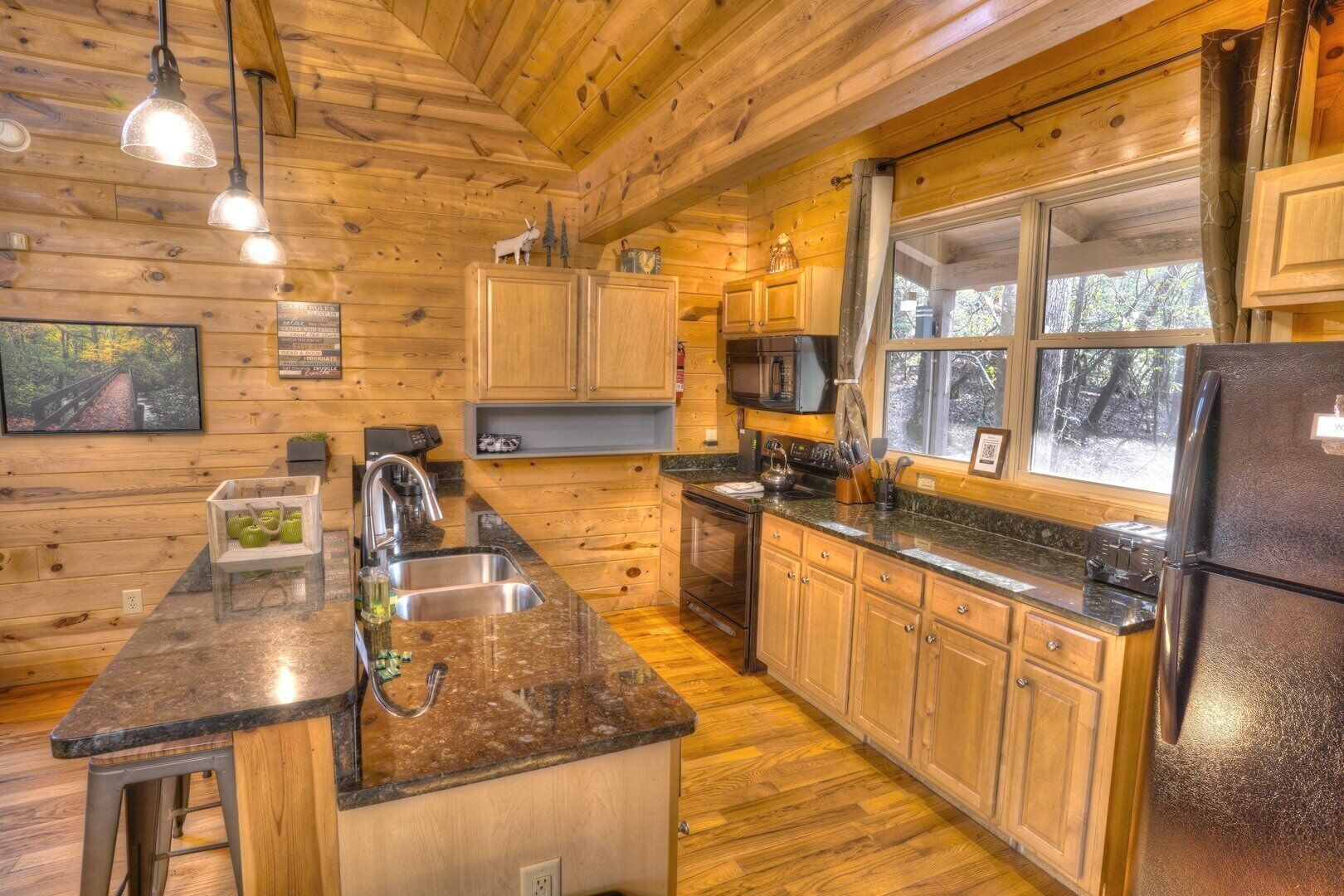 A log cabin kitchen with wooden cabinets, granite countertops, a stainless steel sink, a refrigerator, and a window with a view of trees.