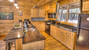 A log cabin kitchen with wooden cabinets, granite countertops, a stainless steel sink, a refrigerator, and a window with a view of trees. - Allure Cabin – Family-Friendly Escape w/ Fire Pit, Creek & Near Helen! (Cleveland)