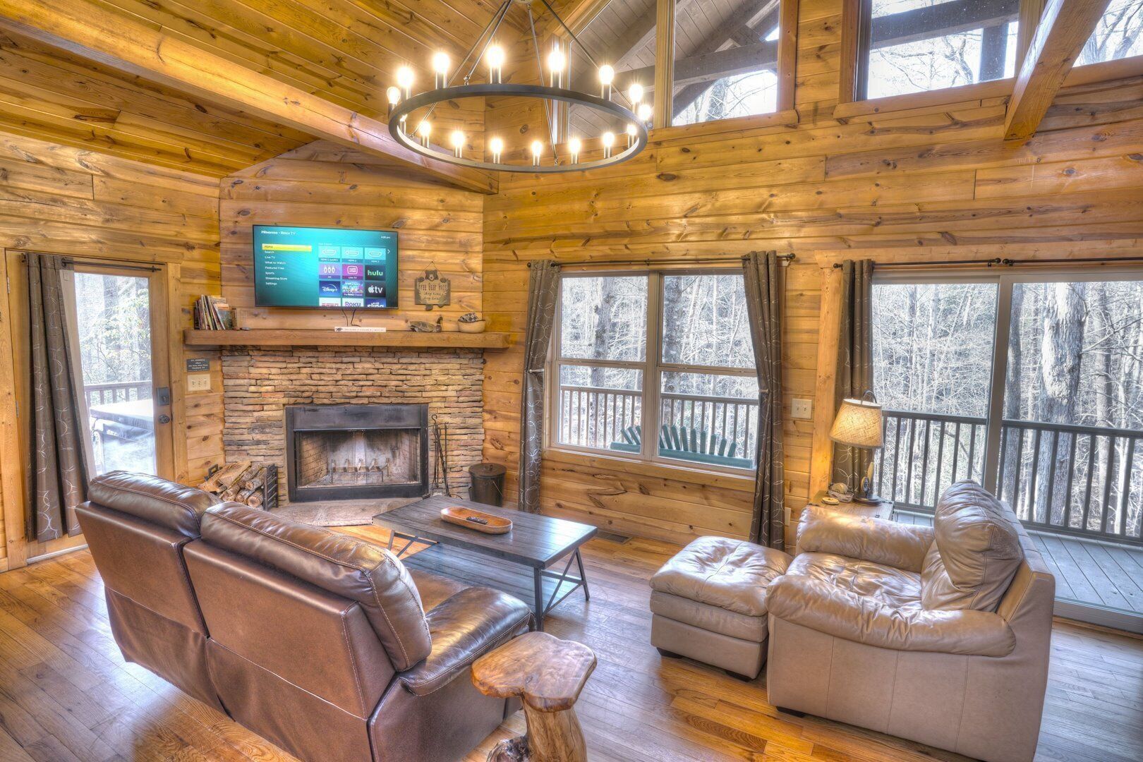 A log cabin interior with a stone fireplace, leather furniture, a television mounted above it, and large windows offering a view of the surrounding forest.