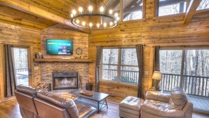 A log cabin interior with a stone fireplace, leather furniture, a television mounted above it, and large windows offering a view of the surrounding forest. - Allure Cabin – Family-Friendly Escape w/ Fire Pit, Creek & Near Helen! (Cleveland)