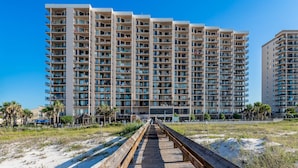Un hotel de gran altura con varios balcones, un paseo marítimo de madera que conduce a la entrada y un cielo azul despejado.