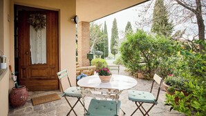 A patio with a round table and four chairs, a wooden door with a wreath, and a garden with various plants and trees. - Villa PoliFlora is a modern apartment in Monticchiello of Pienza in Tuscany. (Monticchiello)