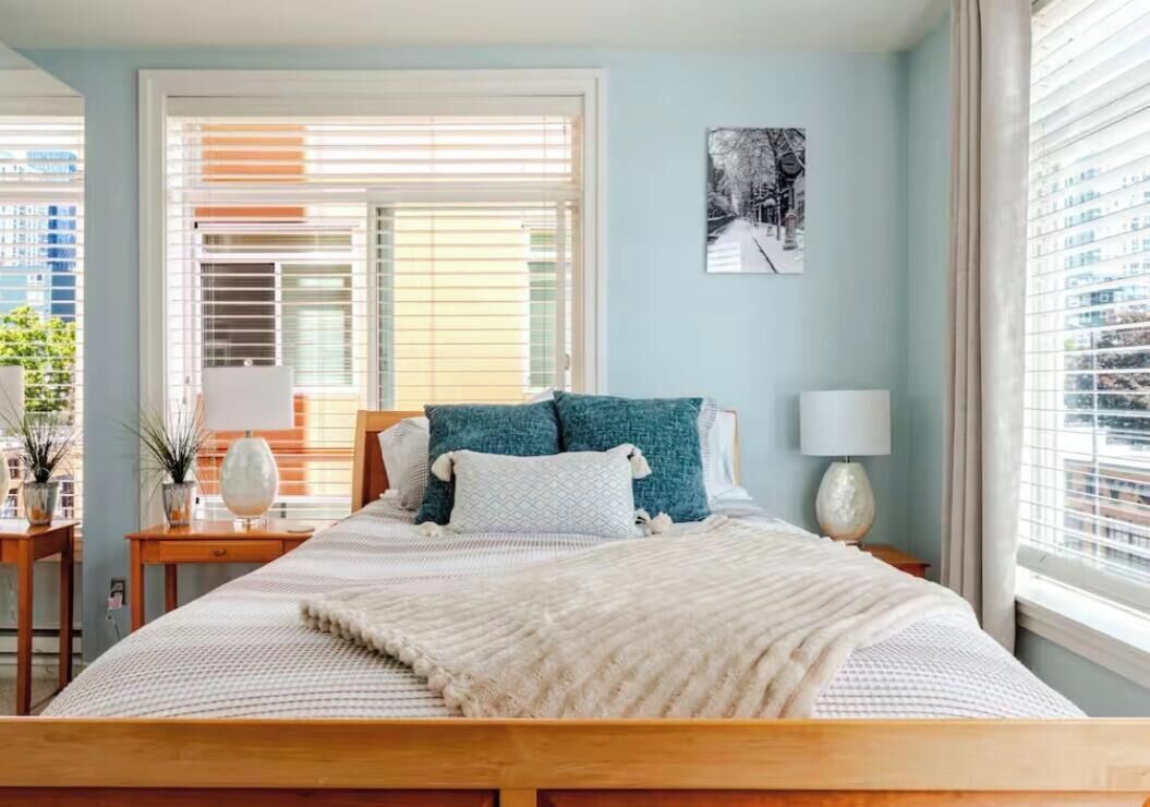 Un dormitorio con una cama grande, dos mesitas de noche, una mesita auxiliar, una lámpara de pared y una ventana con persianas.