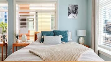 Un dormitorio con una cama grande, dos mesitas de noche, una mesita auxiliar, una lámpara de pared y una ventana con persianas.