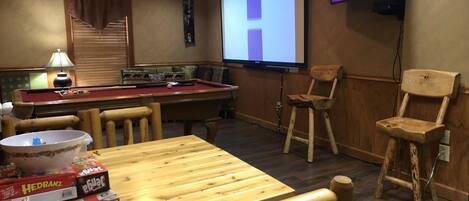 A room with a pool table, wooden chairs, a television mounted on the wall, and a projector screen.