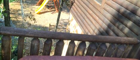 Une terrasse en bois avec une rambarde en métal, donnant sur une aire de jeux avec un toboggan et des balançoires.