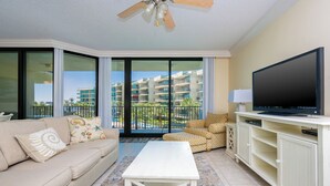 A living room with a sofa, a coffee table, a TV, and a balcony with a view of buildings. - 2 BR 2 BA ~ LOW FLOOR ~ PHOENIX on the Bay 1227! (Orange Beach)