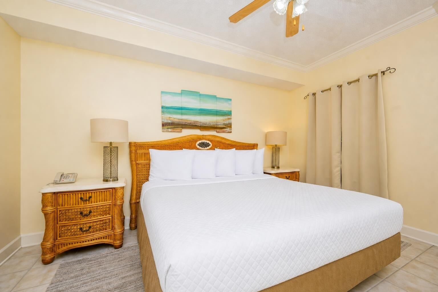 A hotel room with a large bed, wooden nightstands, a painting on the wall, and a ceiling fan.