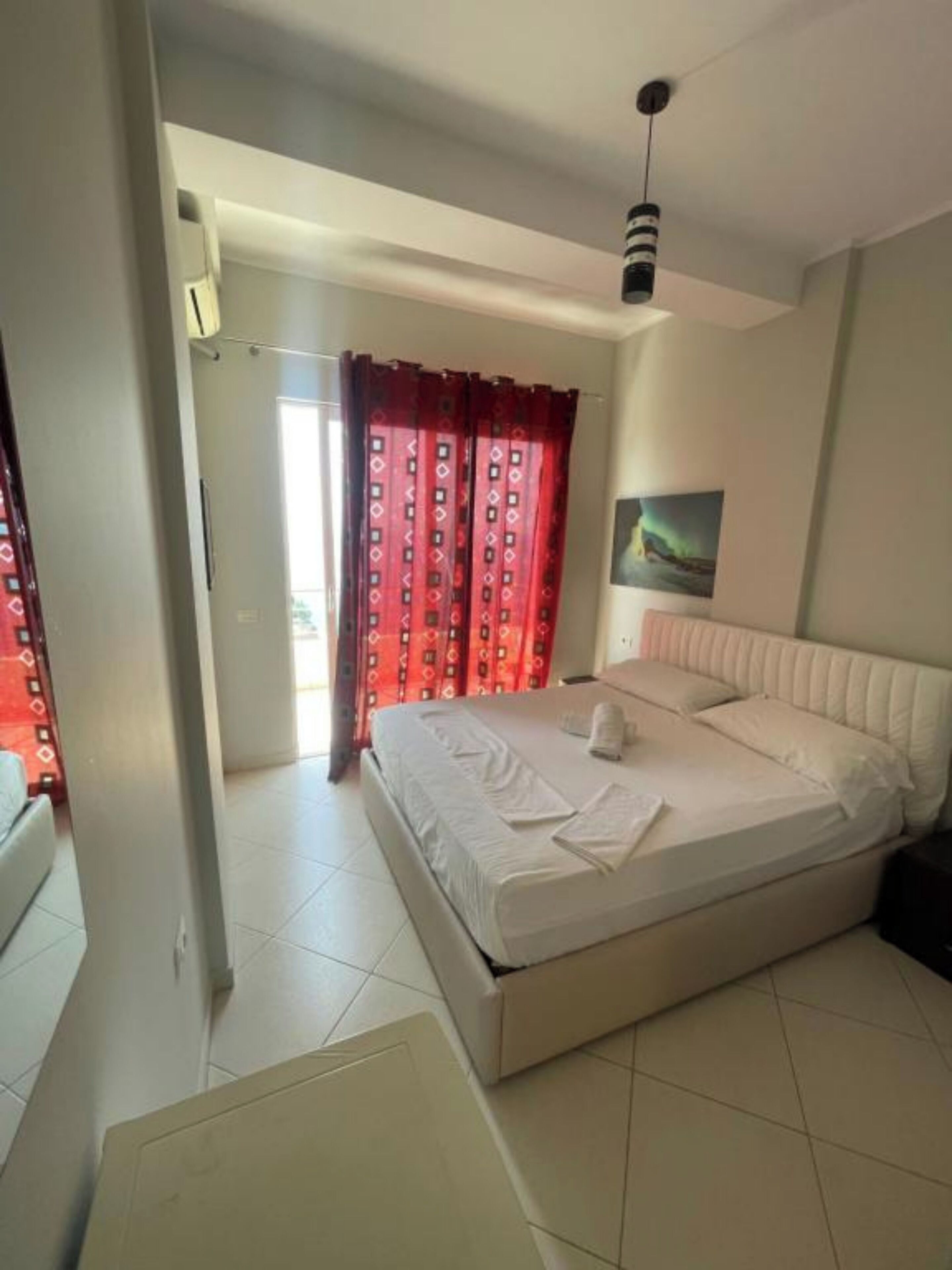 A bedroom with a bed, a painting on the wall, and a window with red curtains.