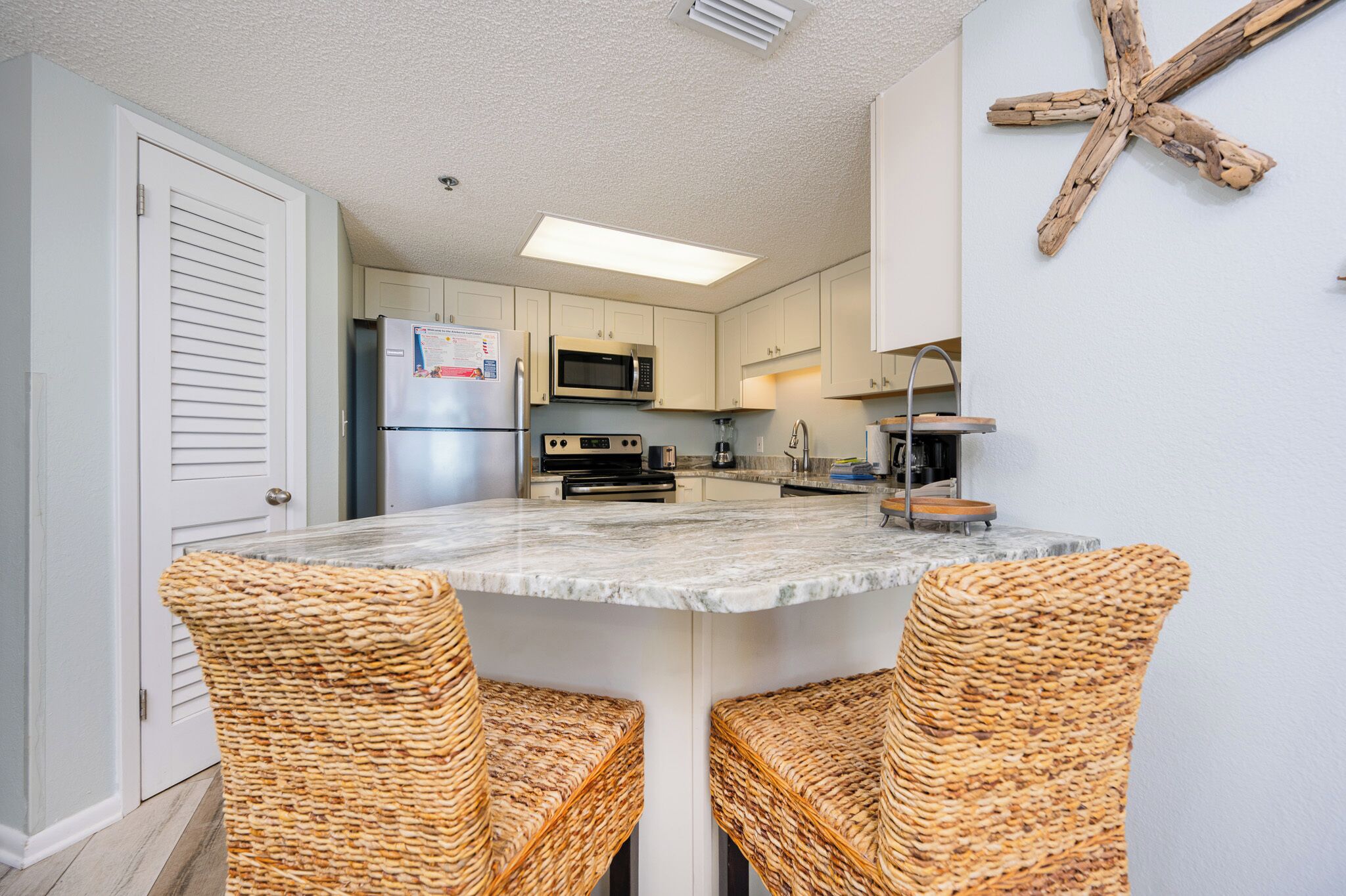 Una cocina con una isla central, dos sillas de mimbre, un microondas, un refrigerador y una decoración de estrella de mar en la pared.