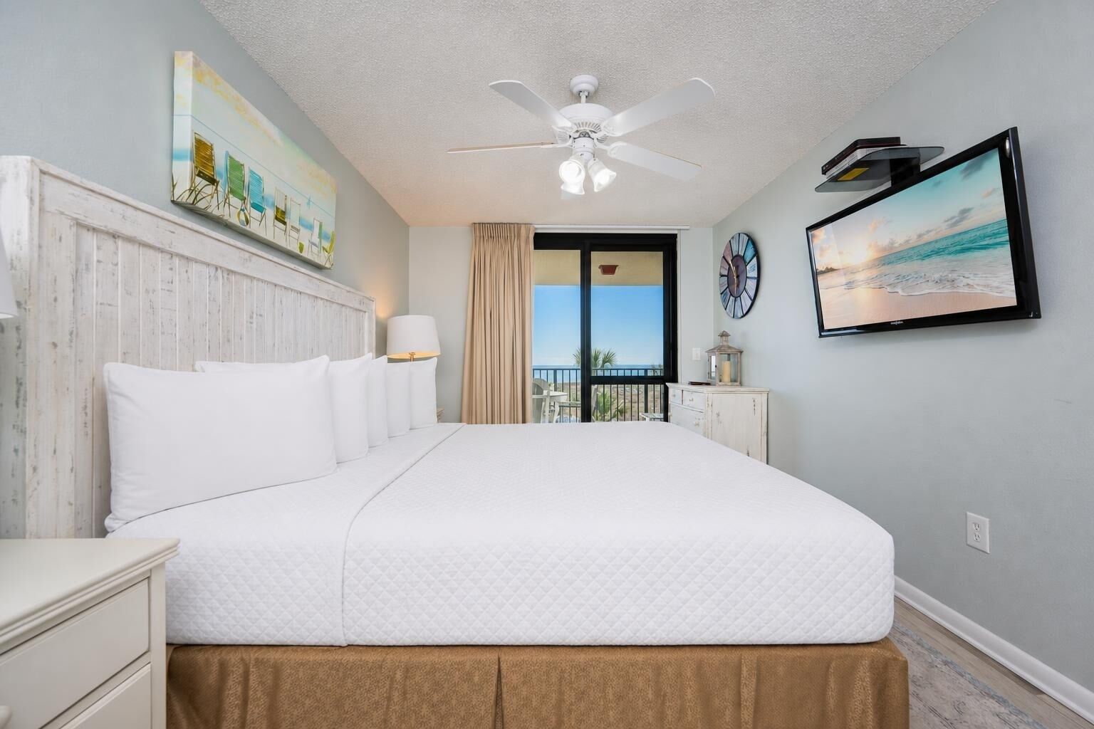 Habitación de hotel con cama, ventilador de techo, vista al océano, un reloj y un cuadro.