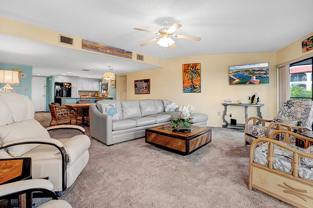 A spacious living room with a grey sofa, a wooden coffee table, and wicker chairs. A ceiling fan is visible, and the room has a light-colored carpet.