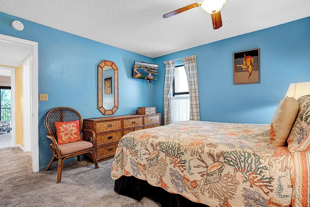 Ein Schlafzimmer mit blauer Wand, einem Bett mit floralem Überwurf, einer Holzkommode, einem Sessel mit Kissen, einem Deckenventilator und einem Fenster mit Vorhängen.