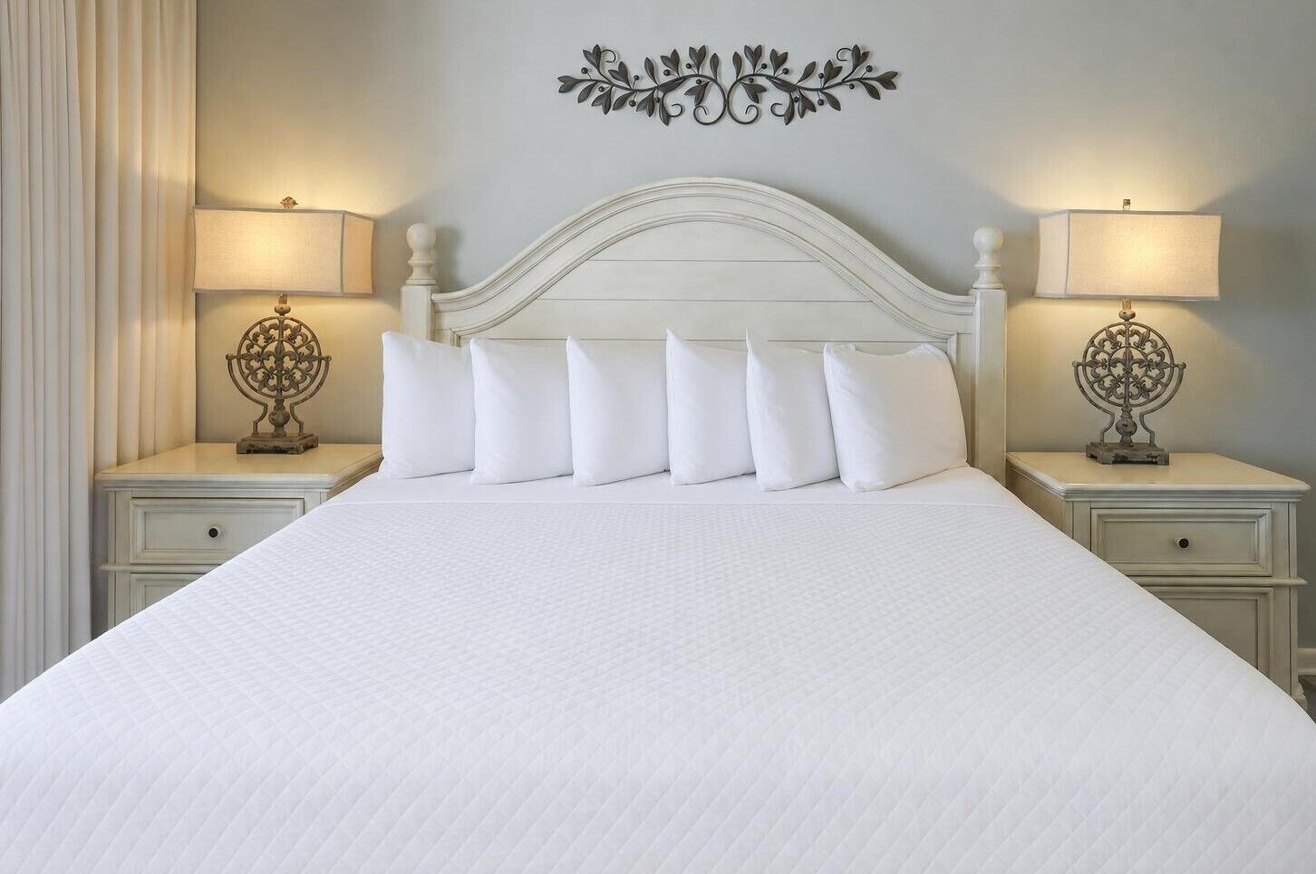 A bed with a white headboard and six pillows, flanked by two bedside tables with lamps and decorative metal stands.