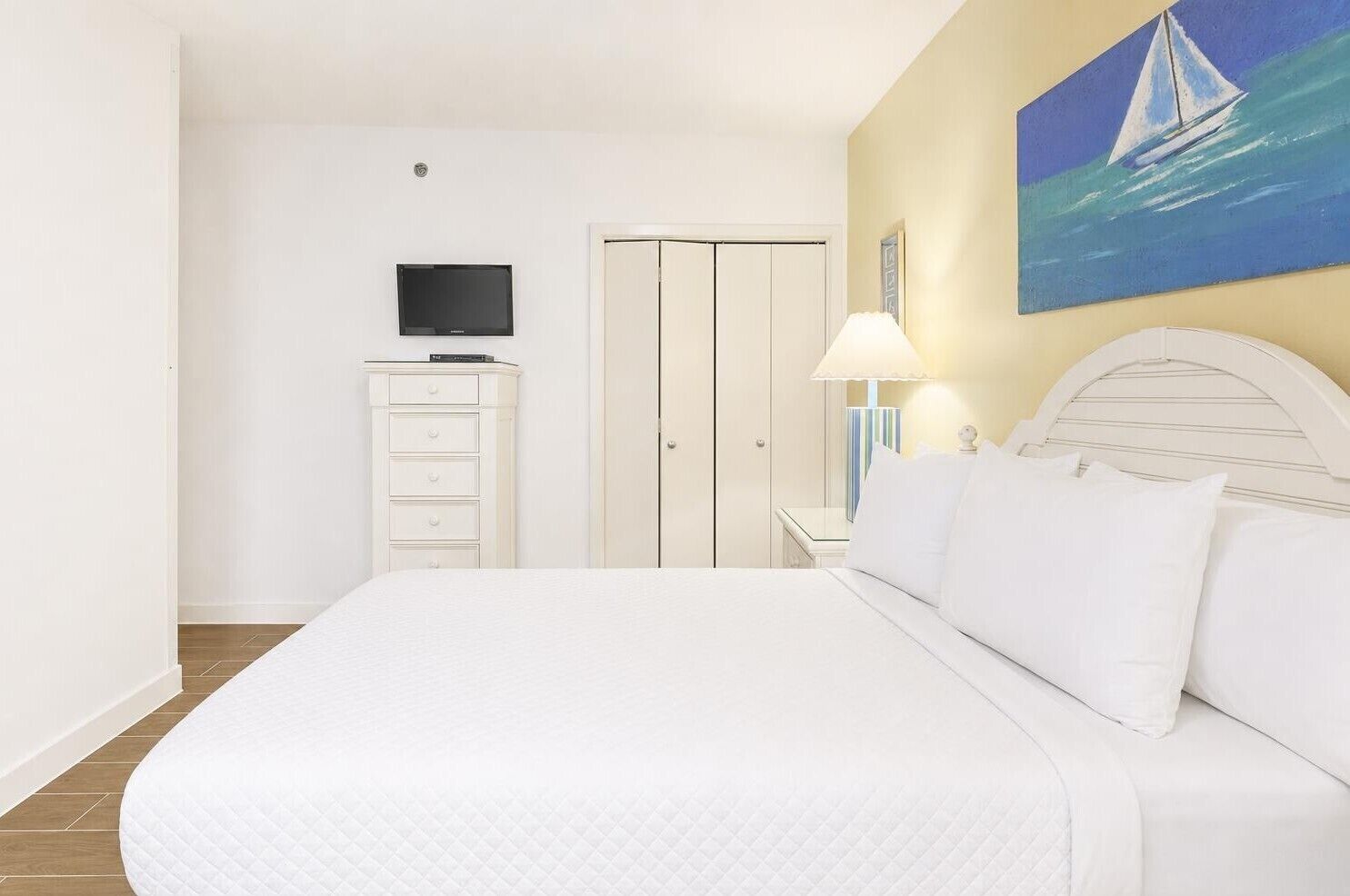 A bedroom with a bed, a TV, a wardrobe, a painting, and a lamp.