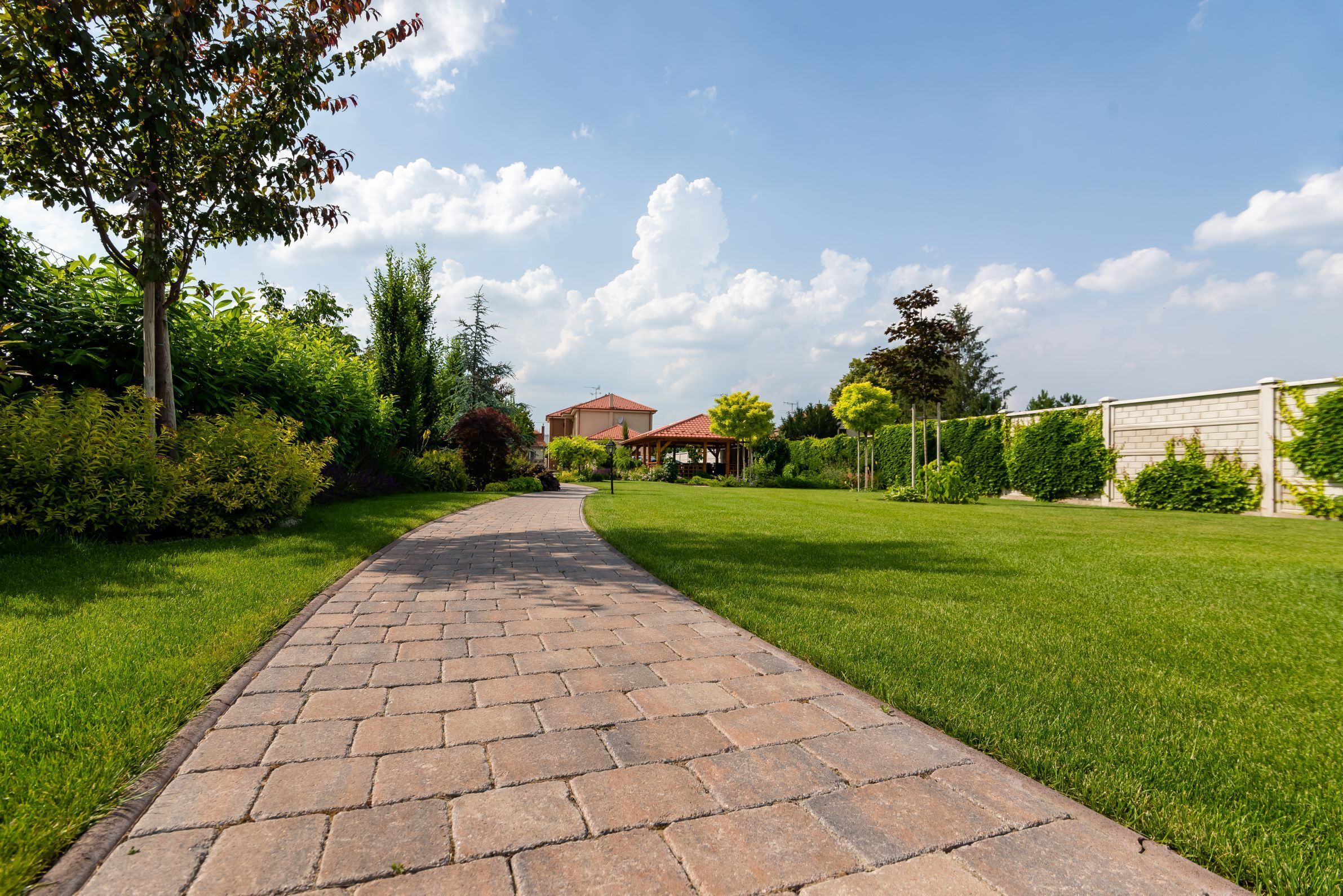 A paved pathway leading to a house with a red roof, surrounded by lush greenery and a clear sky.