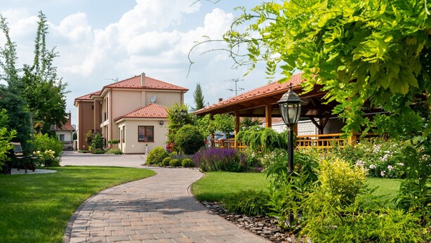 Un chemin pavé mène à un bâtiment dont le toit est recouvert de tuiles rouges, et qui est entouré d’une végétation luxuriante et d’un jardin bien entretenu.