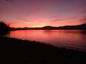 Ein Sonnenuntergang über einem stillen See, im Hintergrund schattenhafte Bäume und Berge.