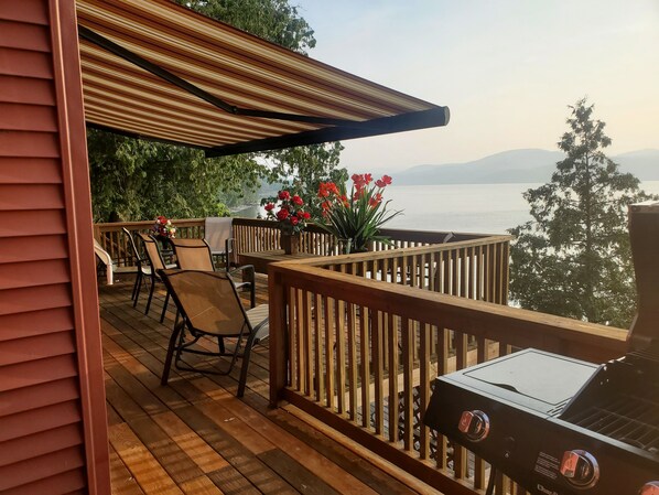 Eine Holzterrasse mit Gartenmöbeln, einem Grill und Blick auf einen See und Berge.