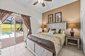 A bedroom with a bed, two bedside lamps, a nightstand, a ceiling fan, and a view of a swimming pool through a sliding glass door.