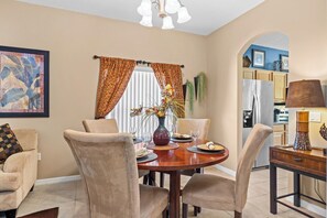 A dining area with a round table, chairs, a vase with flowers, and a kitchen visible through an archway.