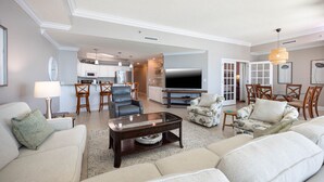A spacious living room with a sofa, armchairs, and a coffee table. There is a kitchen in the background with a TV and a dining area with chairs.