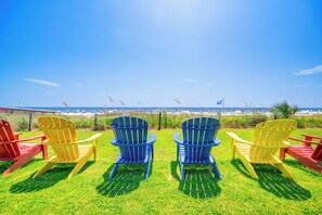 A grassy area with four Adirondack chairs, two blue and two yellow, facing a beach with umbrellas and a clear sky.