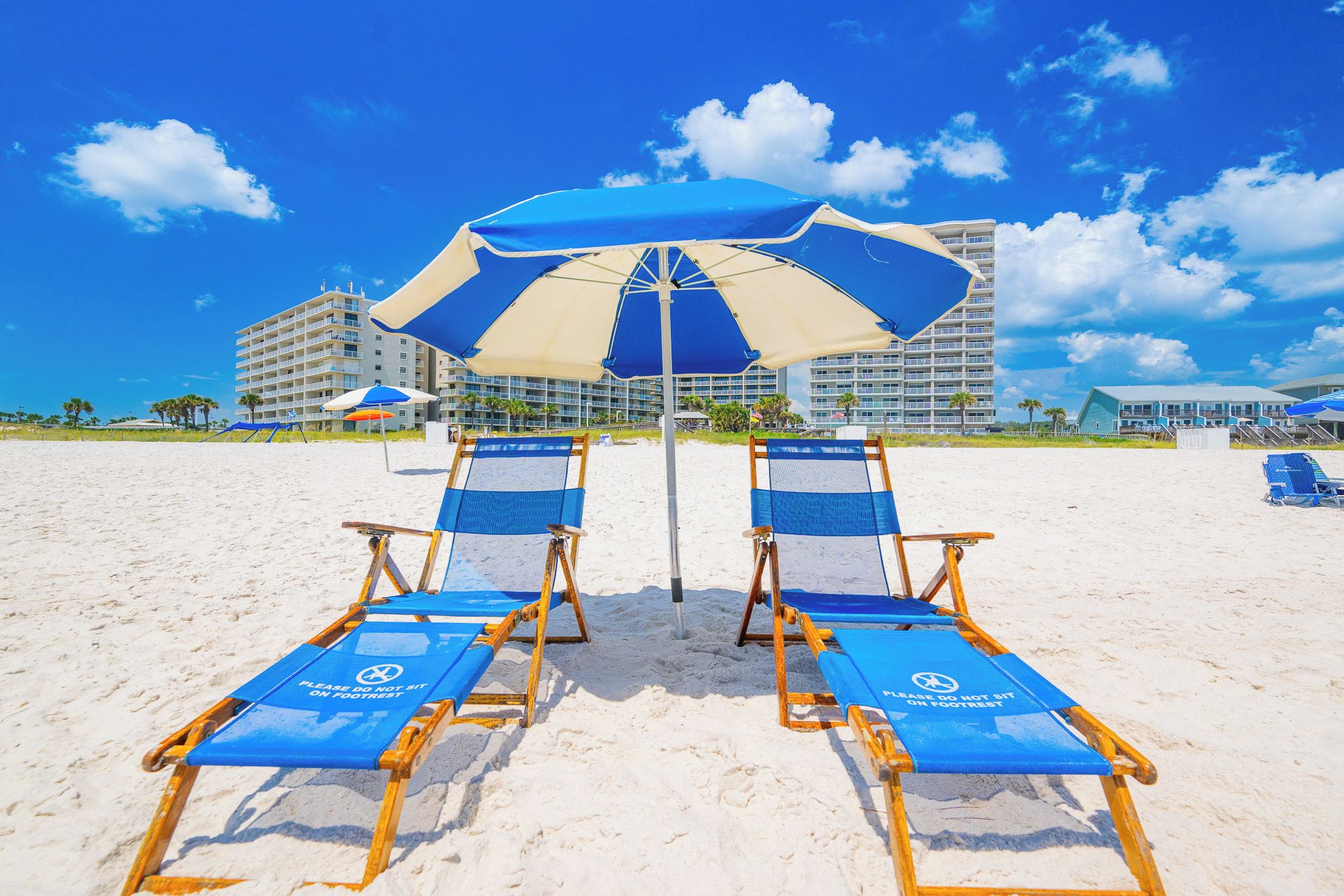 A beach with two lounge chairs under a large umbrella, a sandy beach, and a hotel building in the background.
