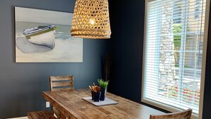 Un coin repas avec une table en bois, des bancs et des chaises. Un luminaire suspendu se trouve au-dessus de la table. Un tableau représentant un bateau est accroché au mur, et une fenêtre avec des stores est visible.