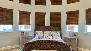 Une chambre avec un grand lit, de nombreuses fenêtres équipées de stores, des tables de chevet avec des lampes et un banc.