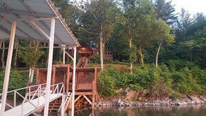 Une cabane en bois dotée d’une terrasse et d’une petite structure en forme de tour, entourée d’une forêt dense et bordée d’une rivière paisible.