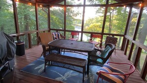Une terrasse extérieure couverte, dotée d’une table et de chaises, d’un ventilateur de plafond, et offrant une vue sur des arbres et une étendue d’eau.