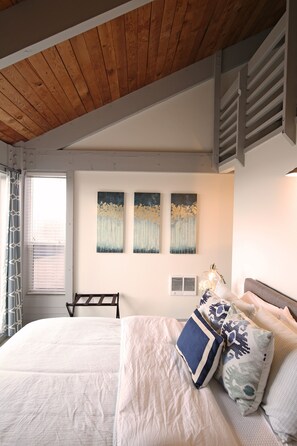 A bedroom with a wooden ceiling, a bed with white bedding and patterned pillows, a window with blinds, and wall-mounted artwork. - Ocean Front...Sand in your toes getaway! (Cloverdale)