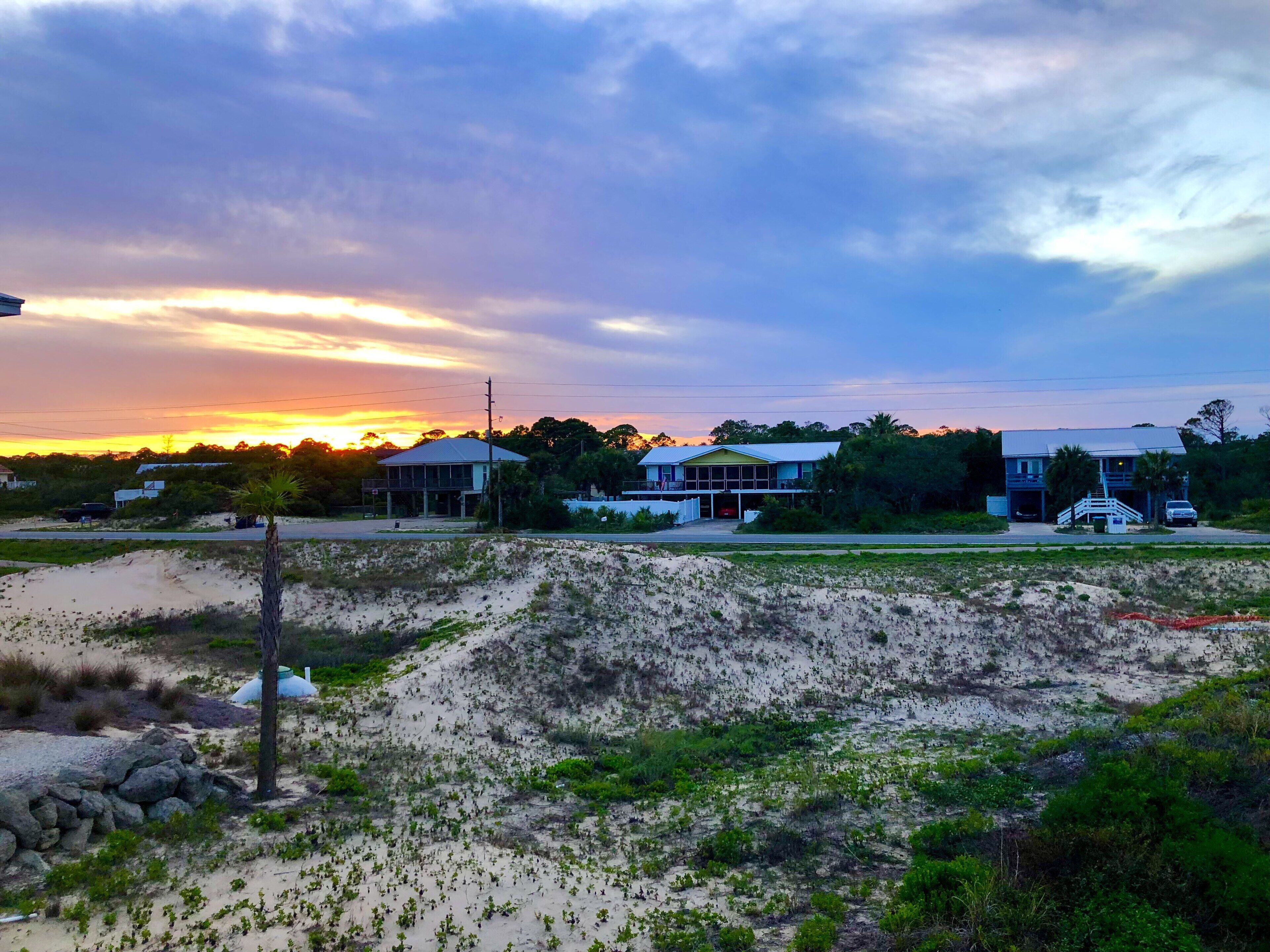 Un paisaje costero con casas, una zona arenosa con vegetación escasa y un atardecer al fondo.