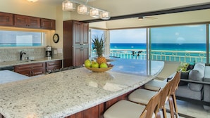 A kitchen with granite countertops, wooden cabinets, and a large island with a fruit bowl. It overlooks a balcony with a view of the ocean. - A View to Die for! 3 bedroom 3 bath on the sand at Waikiki Beach (Honolulu)