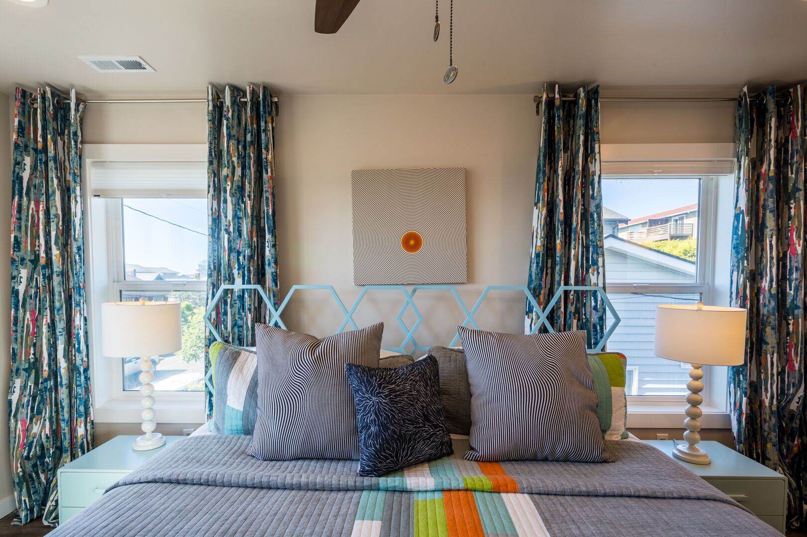 Une chambre à coucher avec un lit, des rideaux à motifs, une œuvre d’art géométrique accrochée au mur et une vue par la fenêtre.