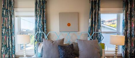 Une chambre à coucher avec un lit, des rideaux à motifs, une œuvre d’art géométrique accrochée au mur et une vue par la fenêtre.