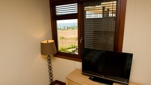 A television on a wooden cabinet, a lamp, and a window with a view of a beach and palm trees.