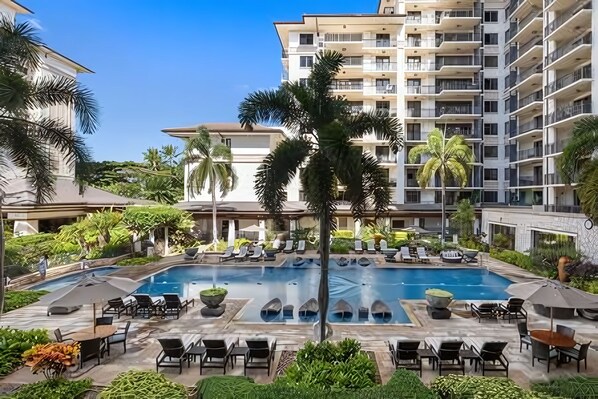 A resort pool area with multiple lounge chairs and umbrellas, surrounded by lush greenery and palm trees.