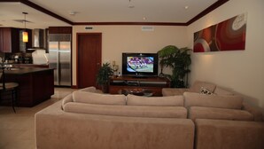 A modern living room with a large beige sectional sofa, a flat-screen TV, a kitchen area with a stainless steel refrigerator, and a dining bar.