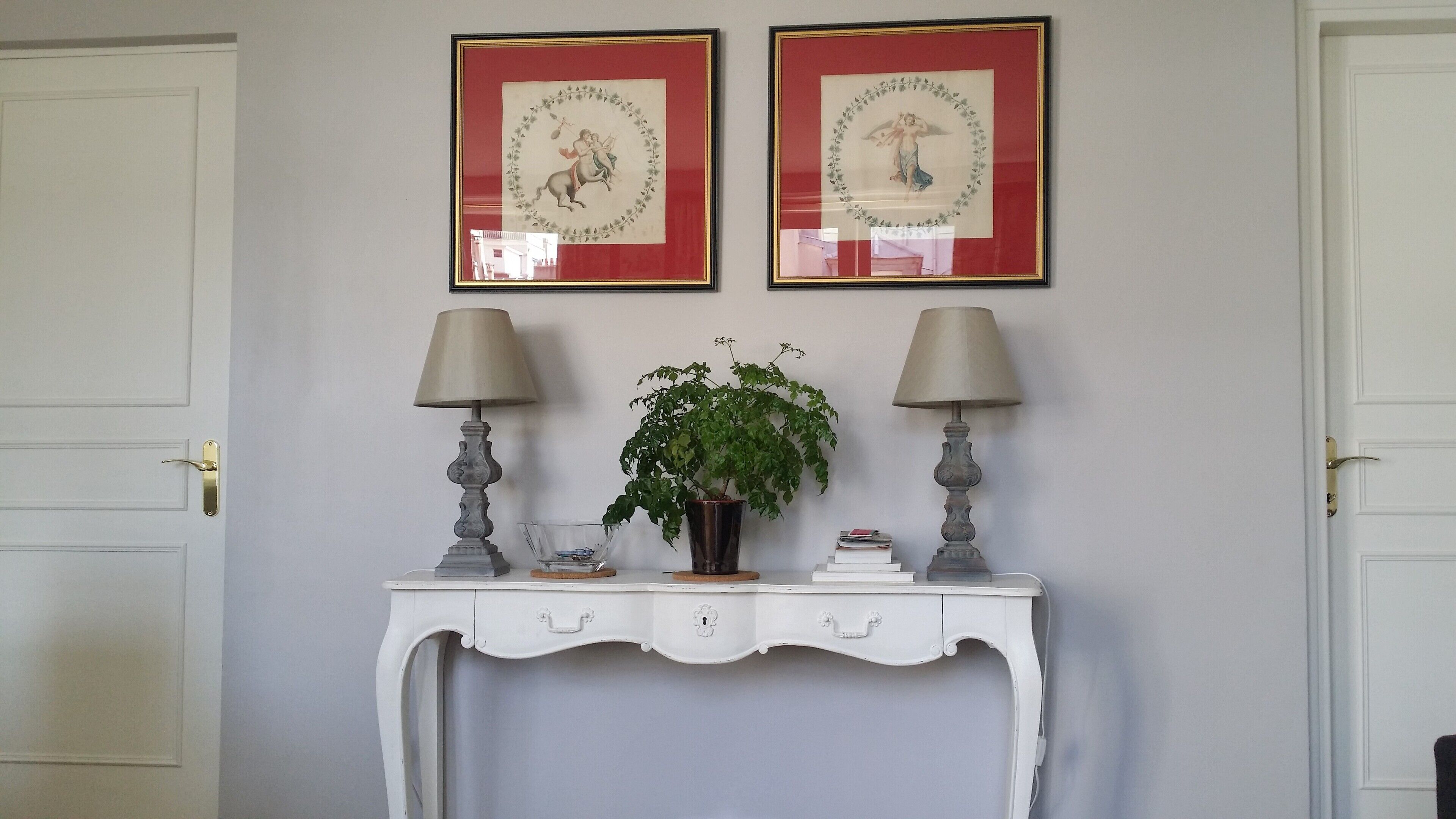 Una consola blanca con dos lámparas, una planta en maceta y dos cuadros en la pared.