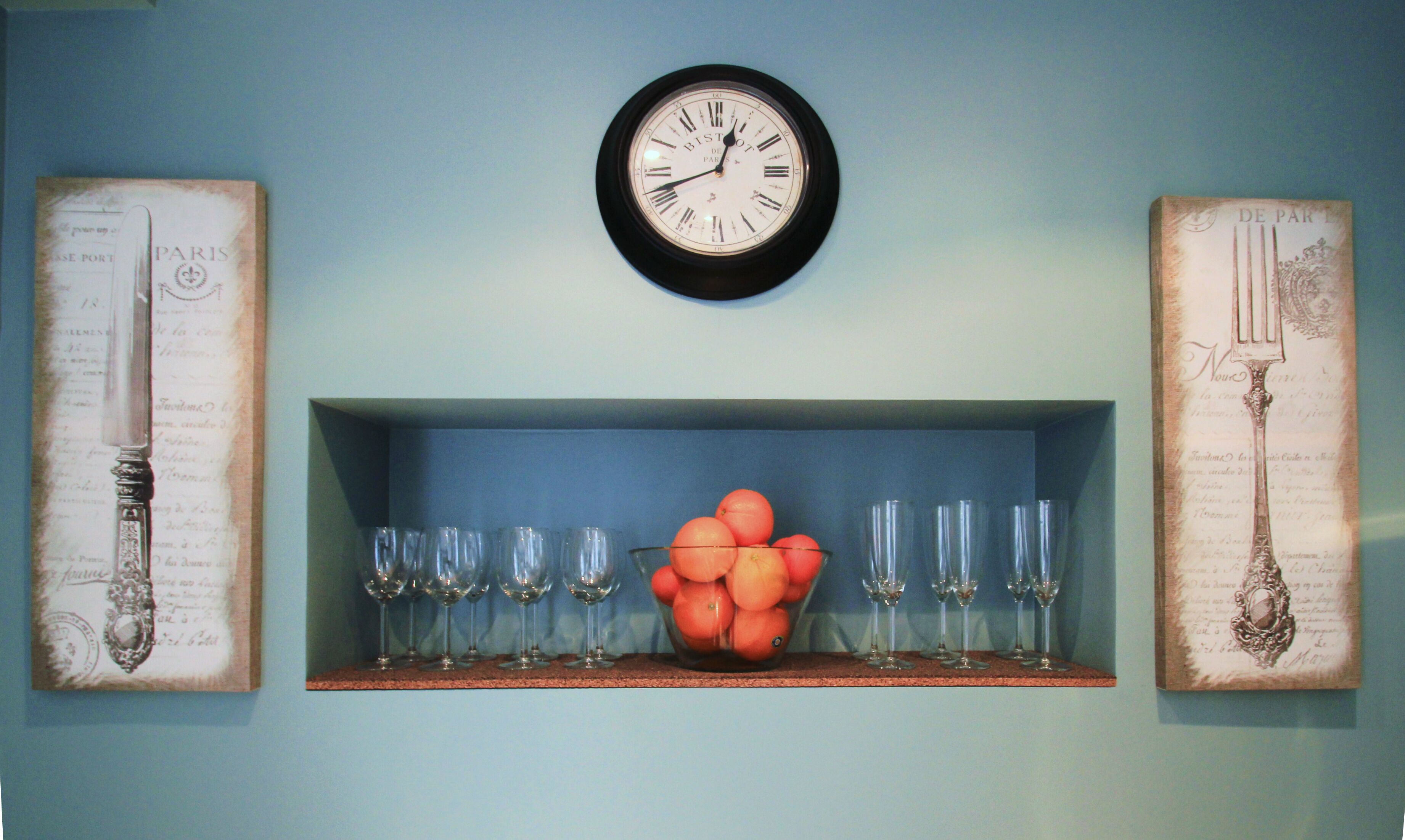 A wall clock above a shelf with wine glasses and a bowl of oranges.