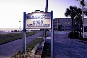 A sign for 'Sommerspell 2396 Scenic Gulf Drive' with a view of a building and palm trees in the background.