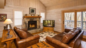 Interior de una cabaña de troncos con sofás de cuero, chimenea de piedra, televisor y mesa de madera.