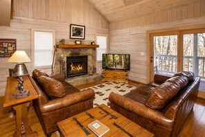 Interior de una cabaña de troncos con sofás de cuero, chimenea de piedra, televisor y mesa de madera.