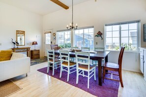 Comedor con una mesa de madera y sillas blancas, una araña y ventanas amplias con vistas al exterior.